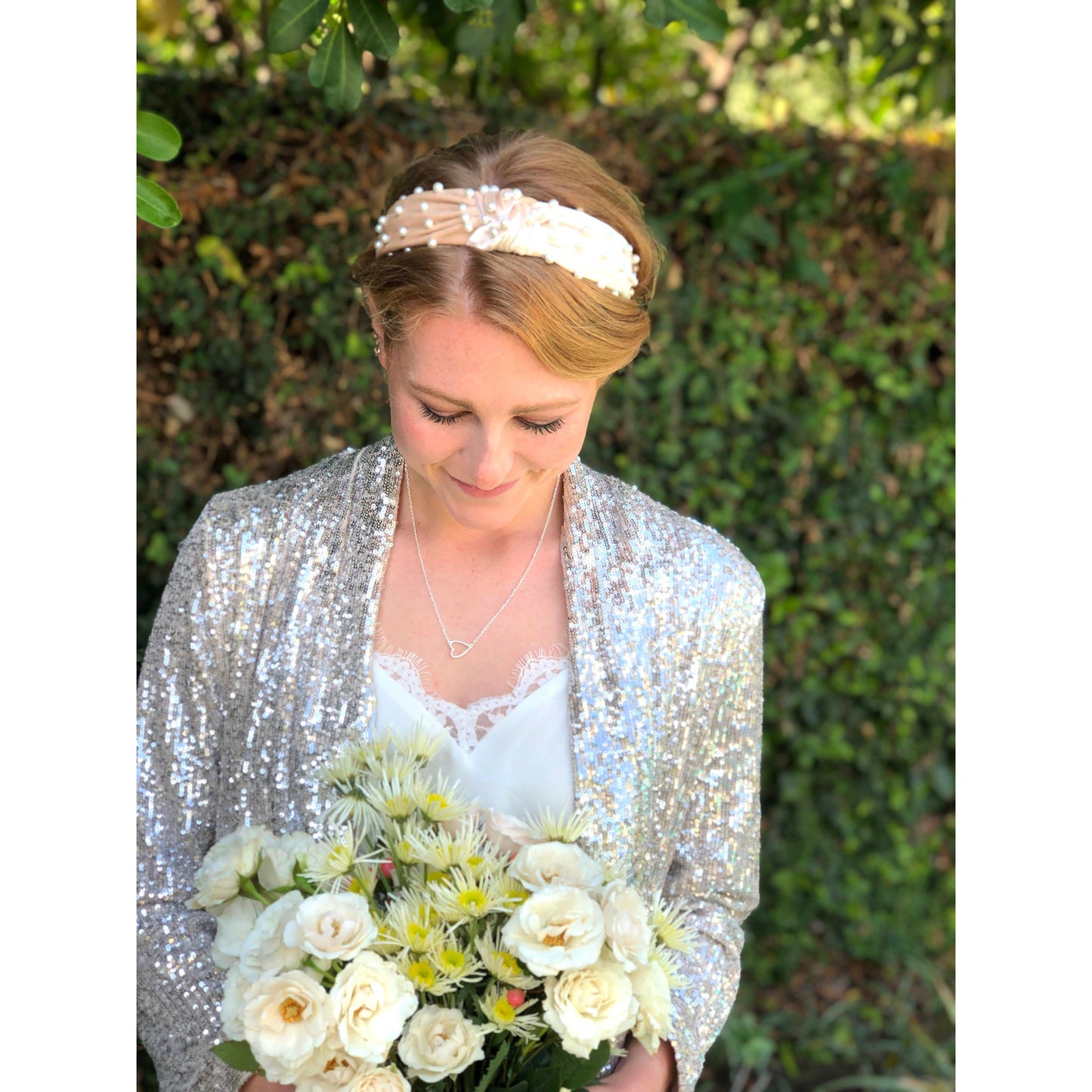 A woman with a pearl headband and sequin jacket holds a bouquet of white and yellow flowers.