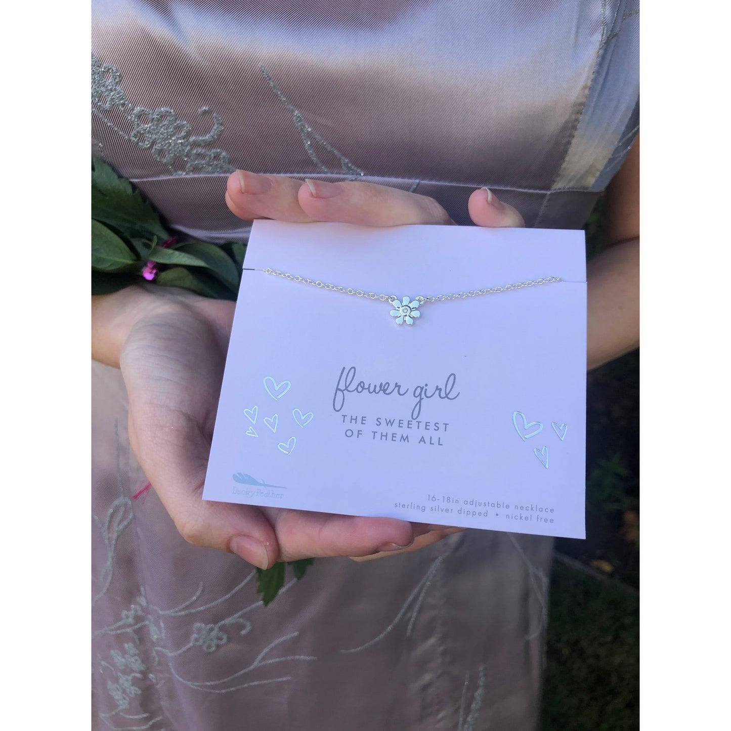 A person holds a "flower girl" necklace with a flower charm on a card that reads "flower girl, the sweetest of them all."