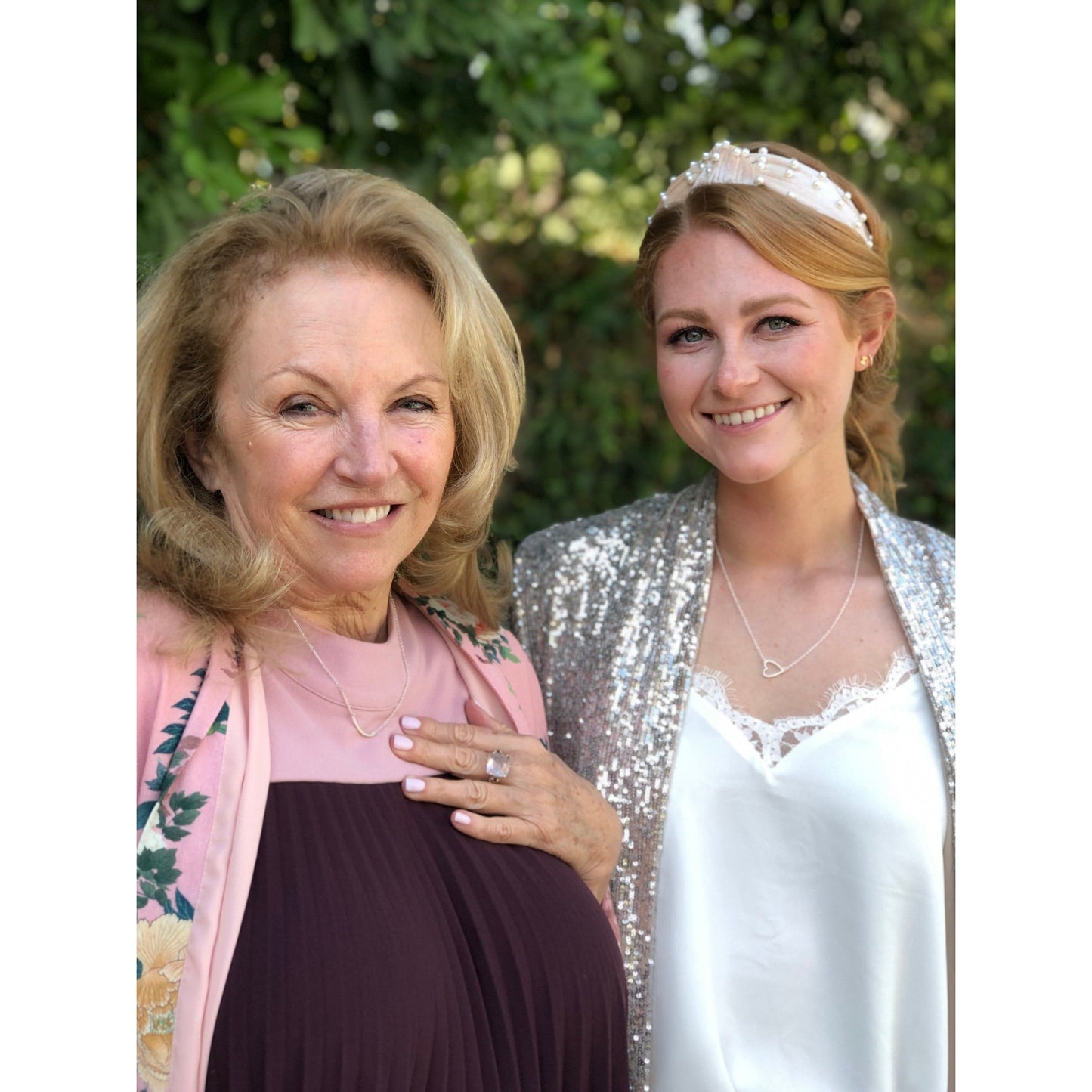 Two women smile, one wearing a sequined jacket and the other a floral-patterned jacket, against a blurred green background.