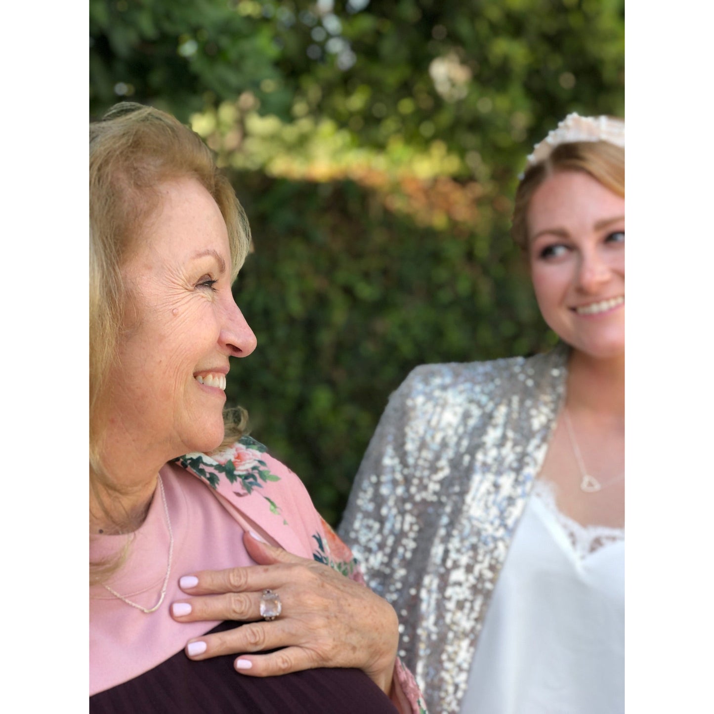 A woman in a floral jacket smiles, with a younger woman in a tiara and sequined jacket smiling in the background.