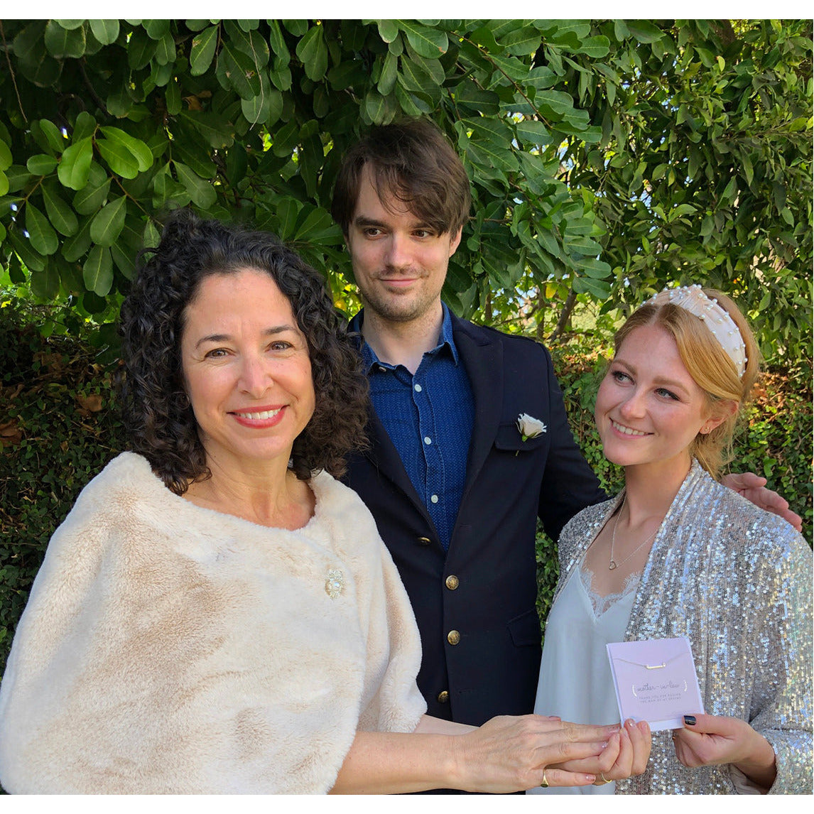 Three people pose outdoors, with a woman in a sparkly jacket holding a necklace card.