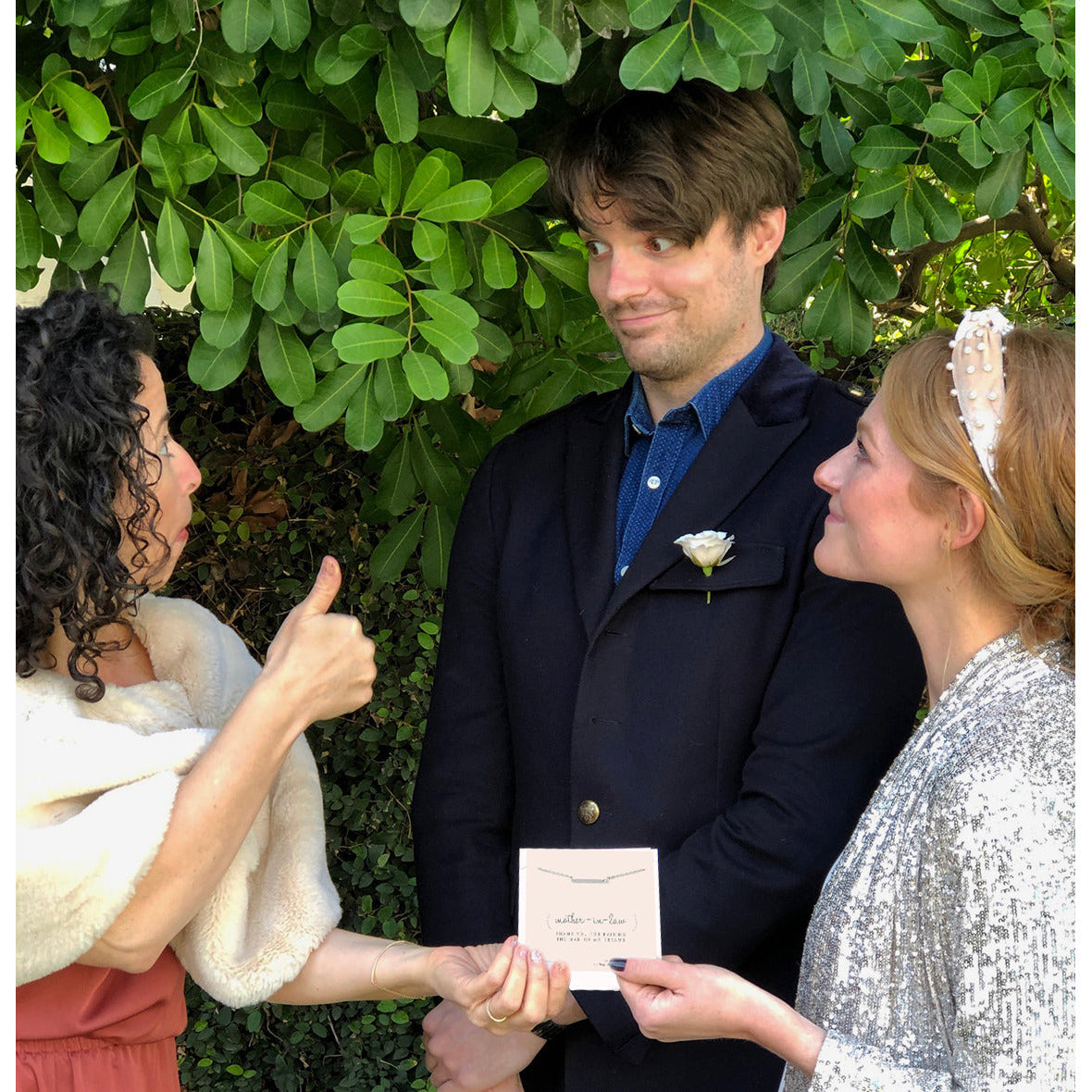 A woman gives a thumbs up while the bride and groom hold a "mother-in-law" gift card near a leafy green backdrop.