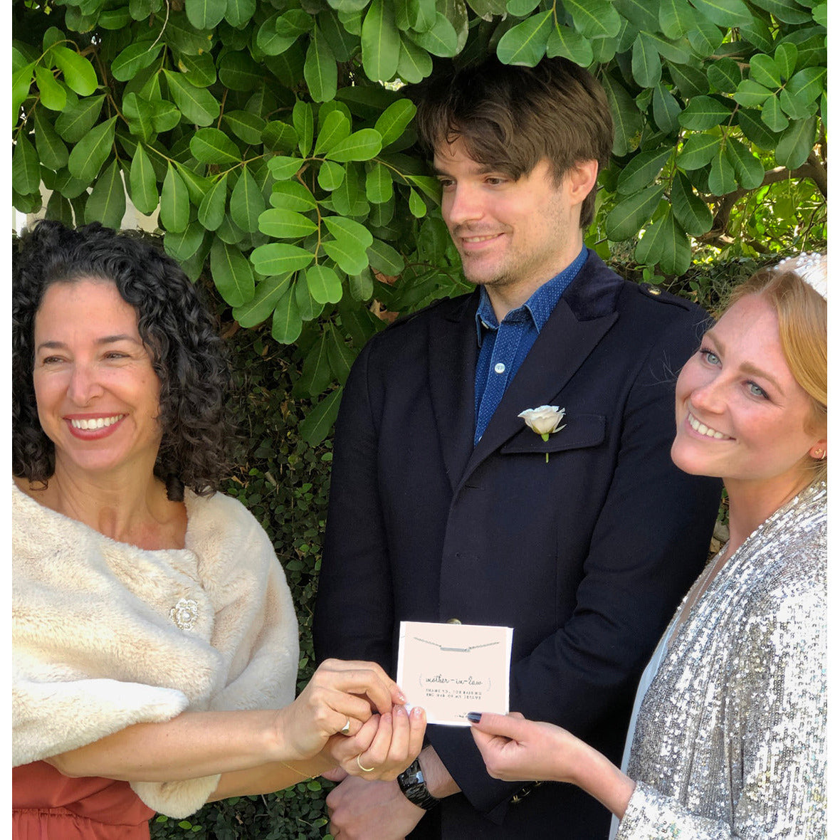 Three smiling people stand in front of a green bush, one holding a card that says "mother-in-law."
