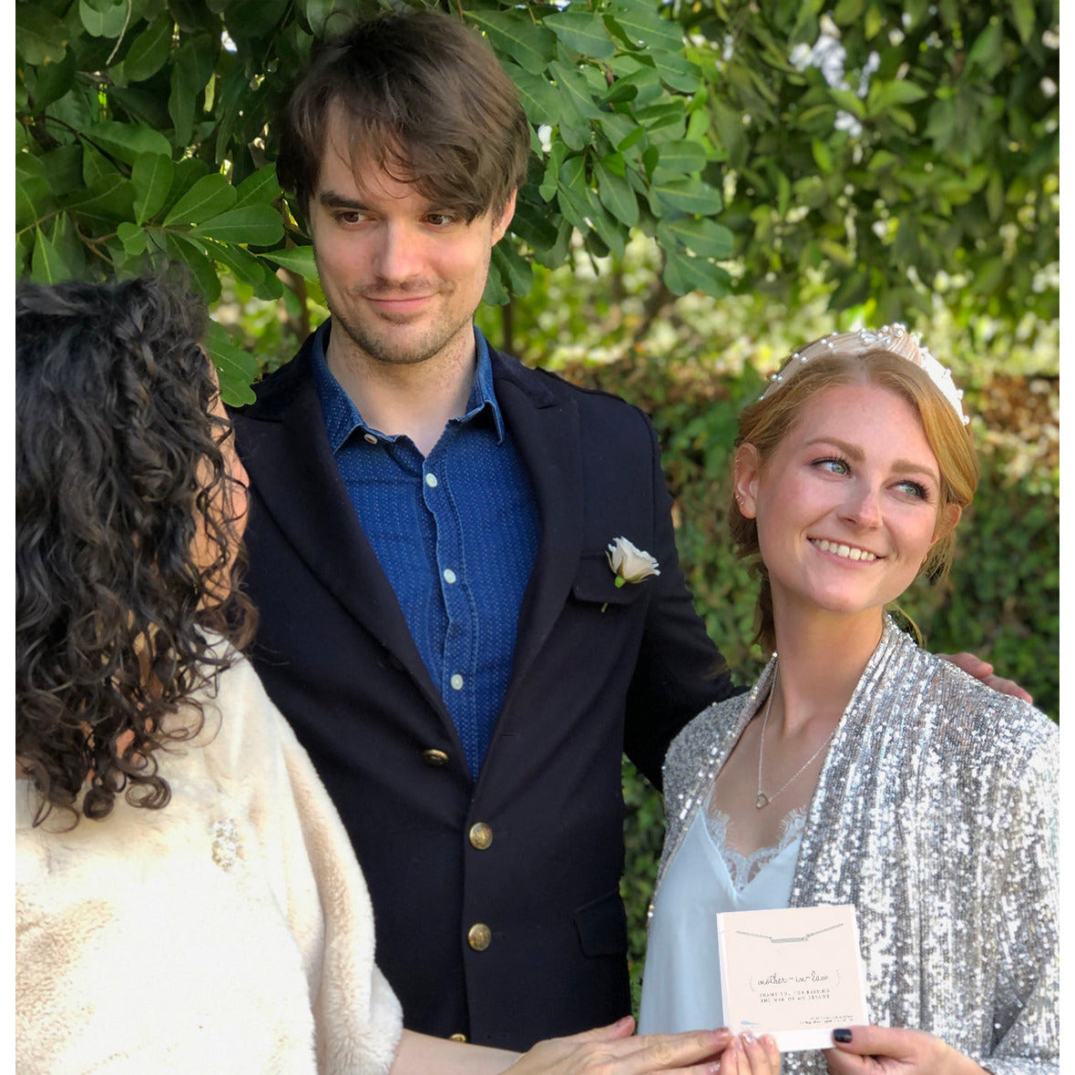 A woman holding a "mother-in-law" gift stands with a man and another woman outdoors.