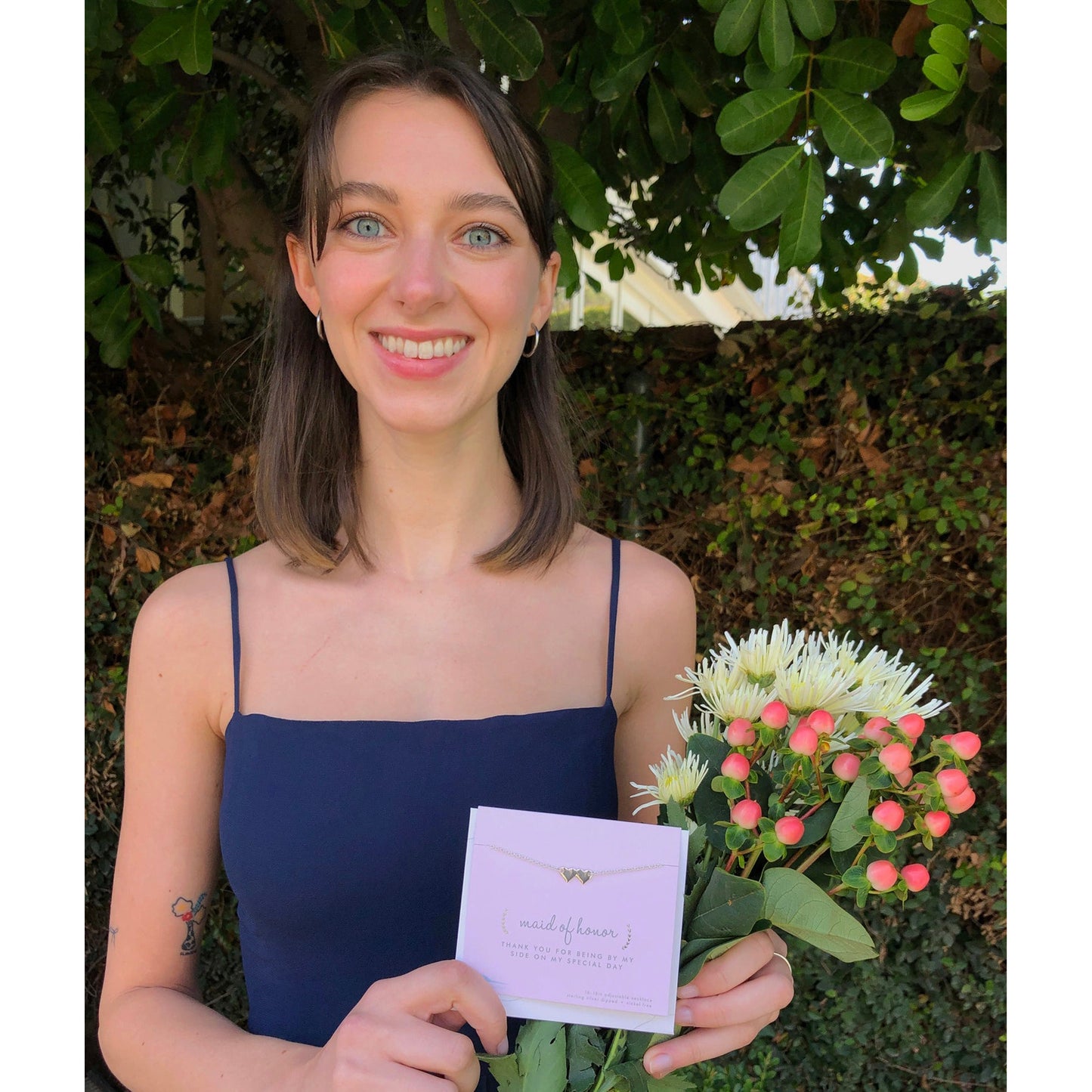 A smiling woman in a navy dress holds a bouquet of white and pink flowers and a "Maid of Honor" thank you card.