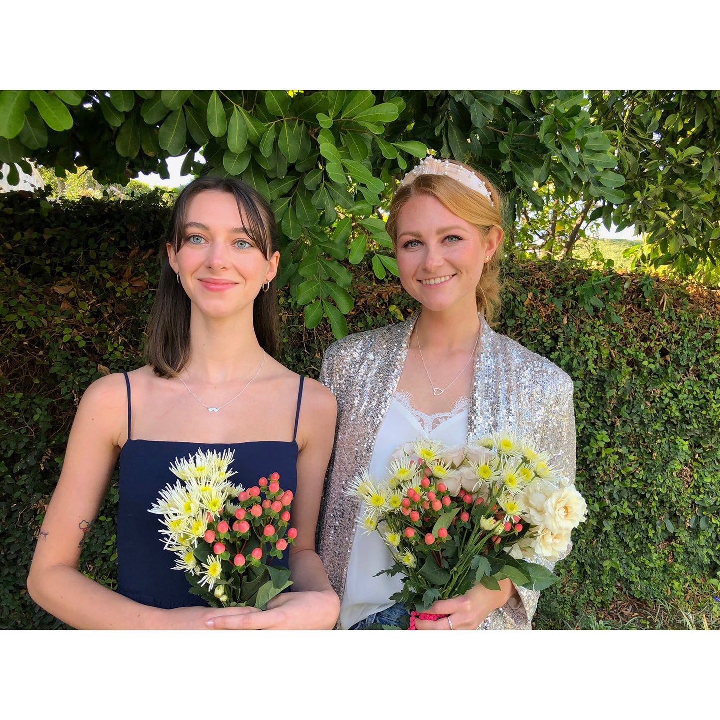Two women stand outdoors holding bouquets.