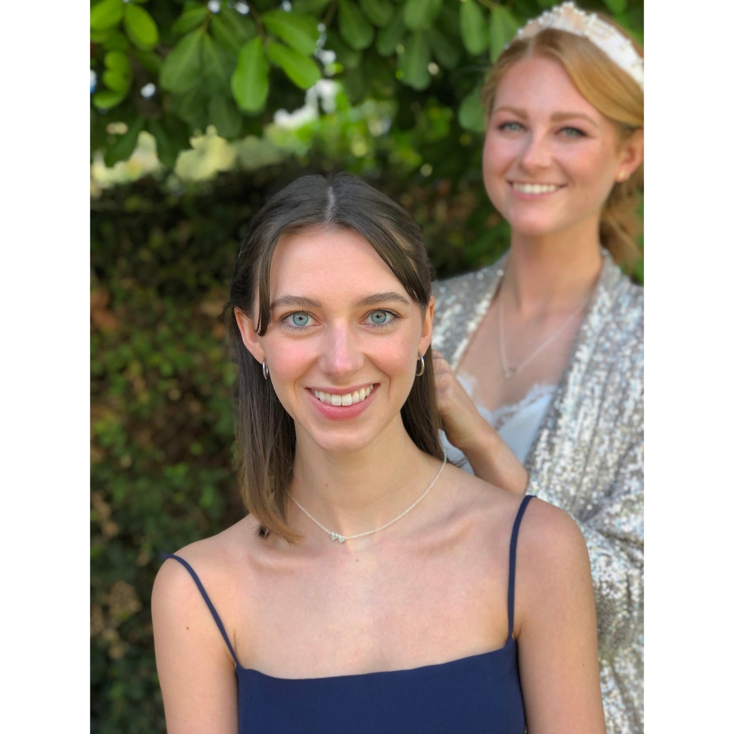 Two smiling women stand near a leafy green background.