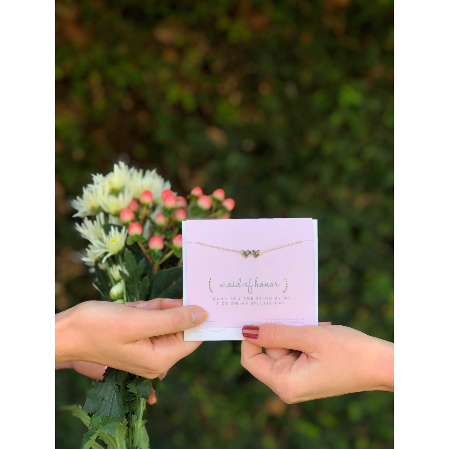 Two hands hold a "maid of honor" card and a bouquet of flowers.