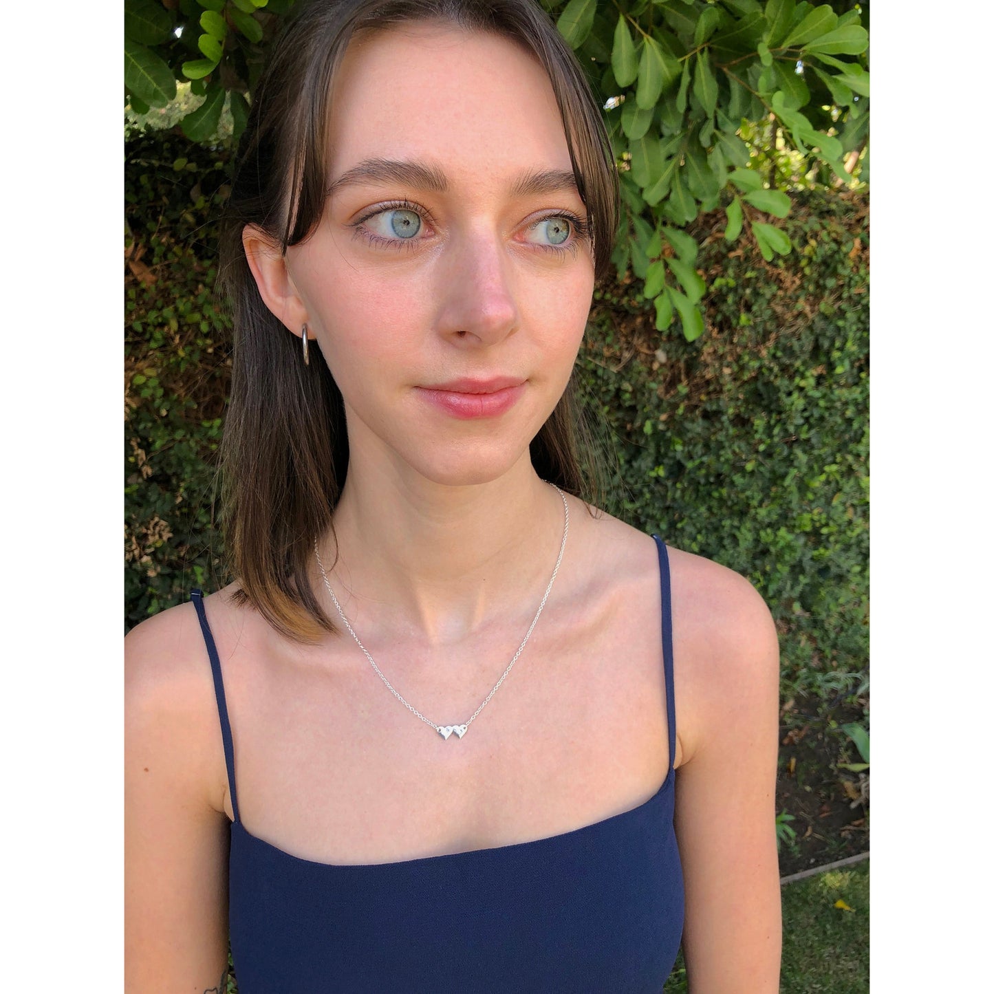 A woman with blue eyes and a navy blue dress poses in front of green foliage, wearing a silver necklace with two small heart pendants.