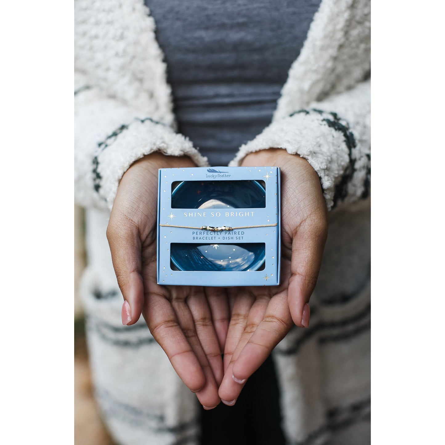 A person holding a light blue gift box set with the words "SHINE SO BRIGHT" in their hands.
