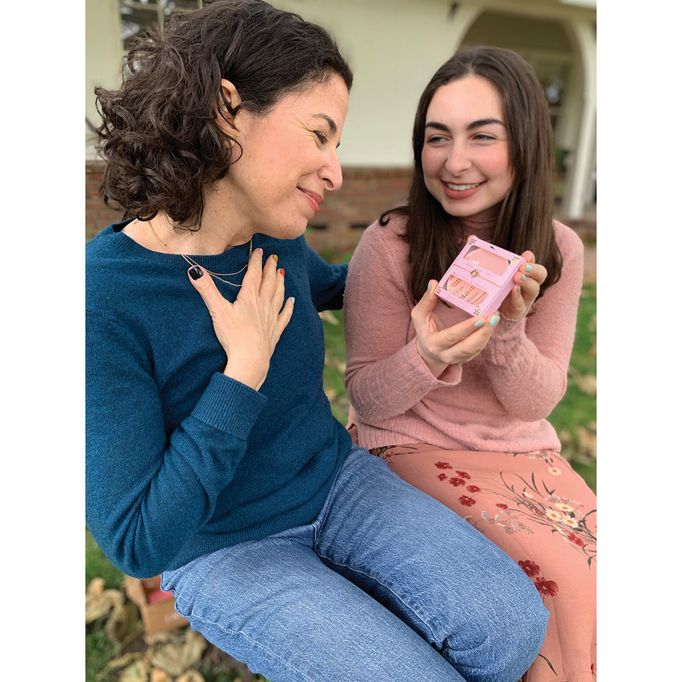 Two women sit together outside, one holding a pink gift and looking at the other with a smile.