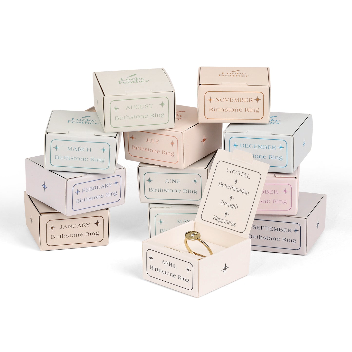 A stack of "Lucky Feather" Birthstone Ring boxes, one open to reveal a gold ring with a crystal.