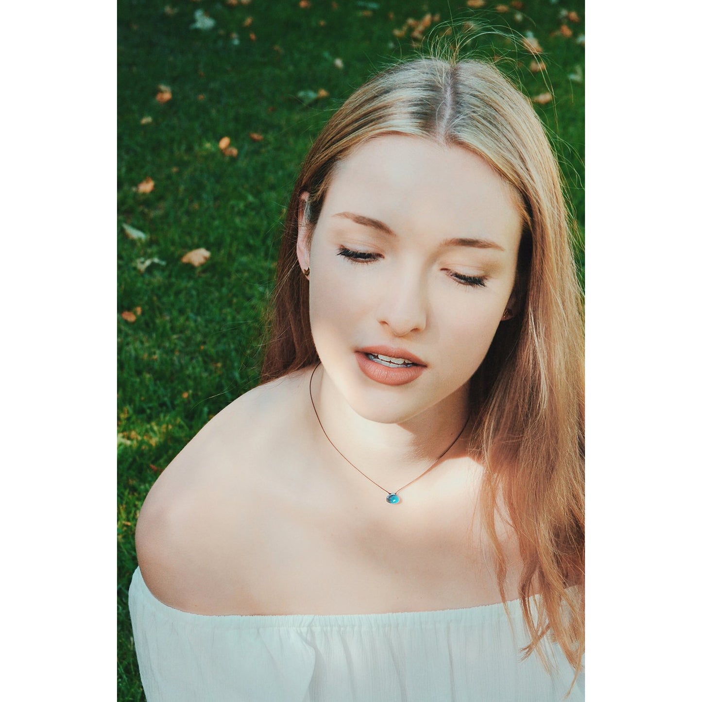 A woman with long blond hair and a blue necklace looks down with her eyes closed.