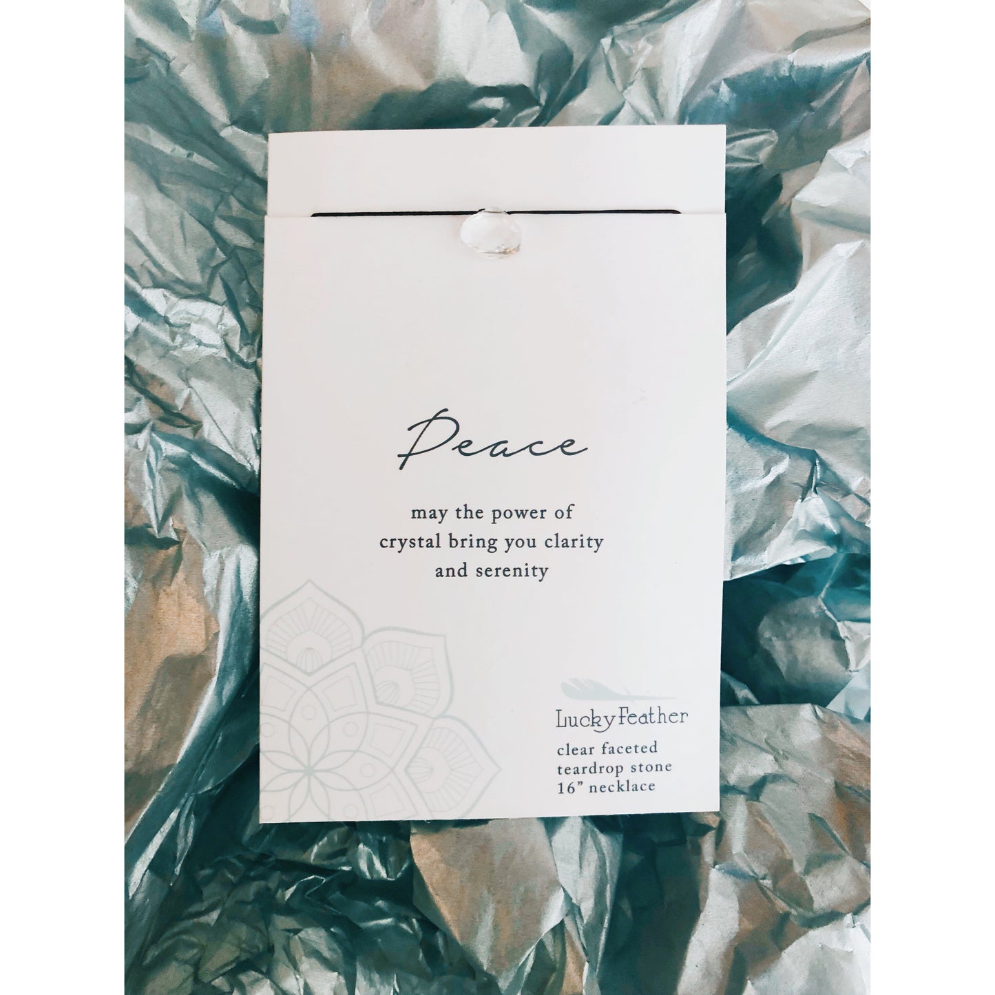 A "Peace" necklace by Lucky Feather rests on crinkled, shiny teal tissue paper.