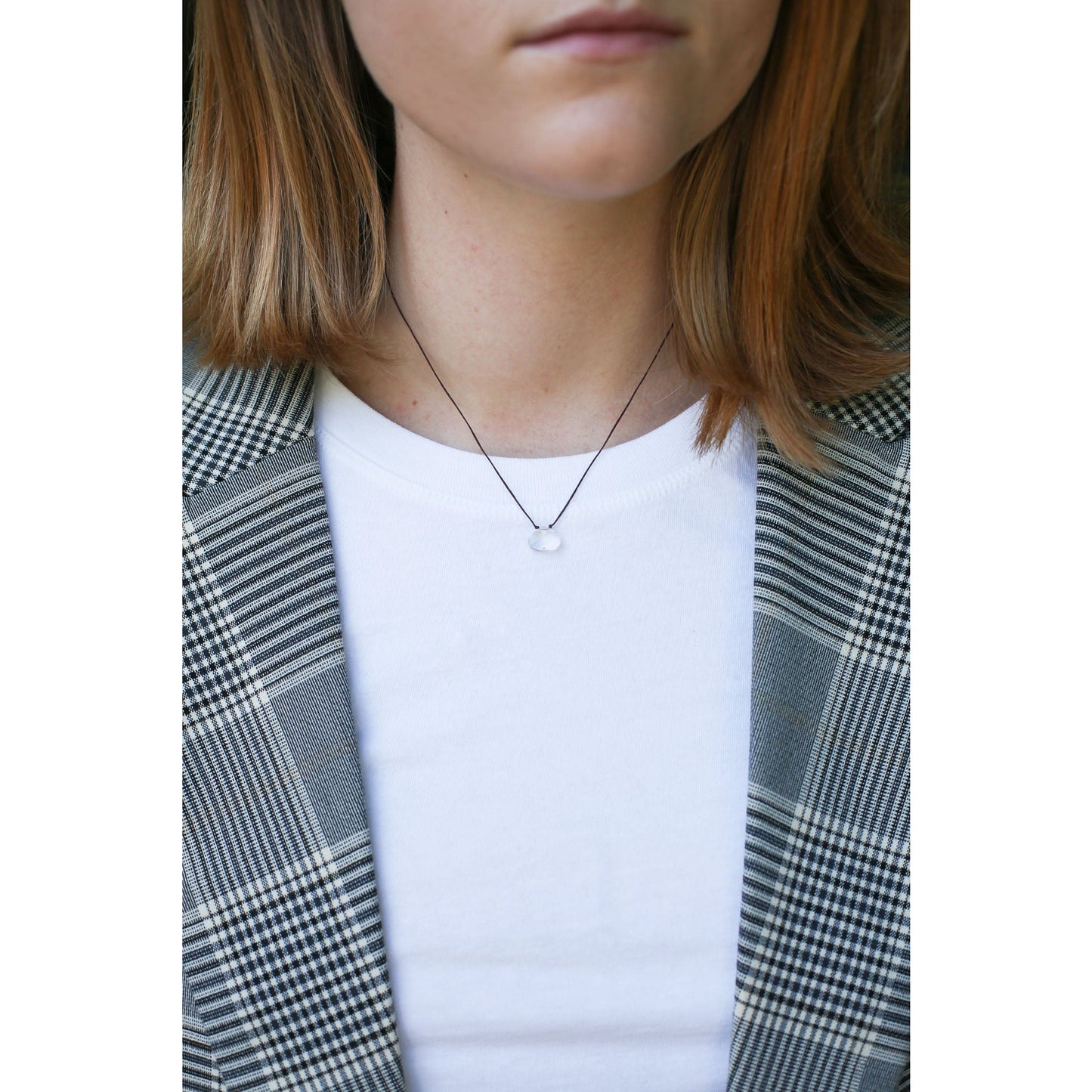A person wears a clear pendant necklace, a white t-shirt, and a gray plaid blazer.