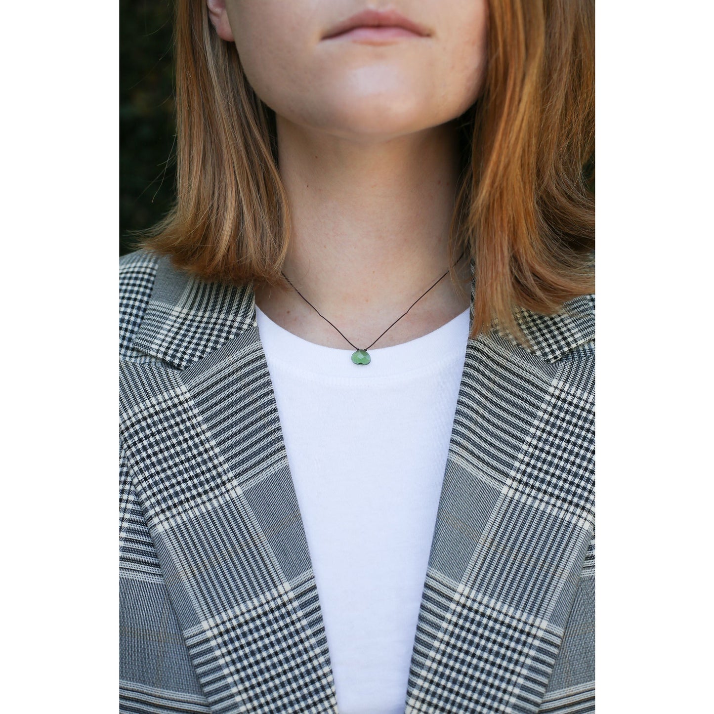 A person wears a black cord necklace with a small, round, green pendant, a white t-shirt, and a gray and white plaid blazer.