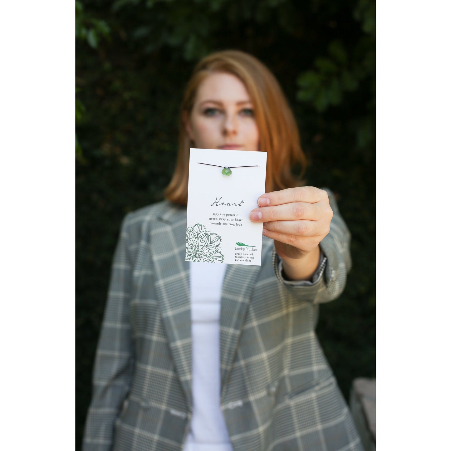 A woman in a plaid blazer holds up a card displaying a green teardrop pendant necklace.