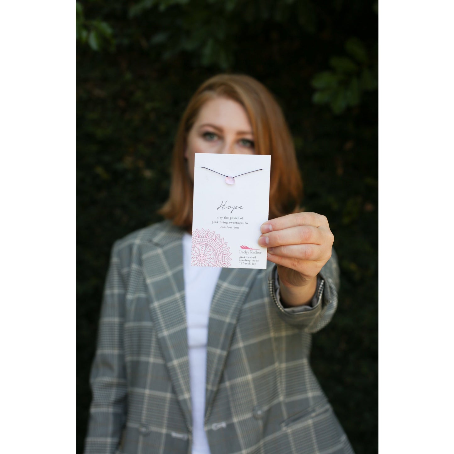 A person wearing a plaid blazer holds up a jewelry card for a "Hope" necklace with a pink teardrop stone.