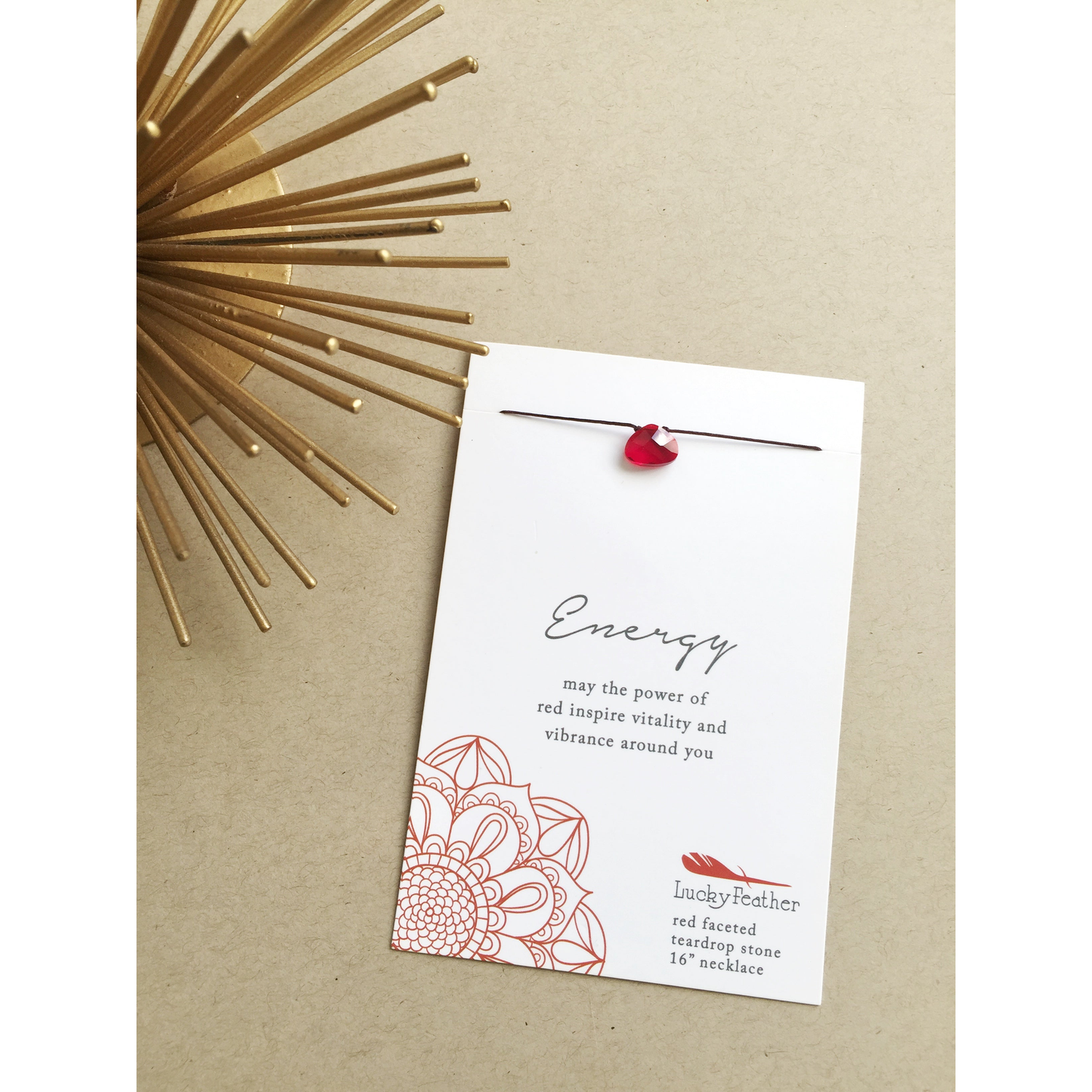 A red faceted teardrop stone necklace on a card with the word "Energy" on it, next to a gold decorative piece.