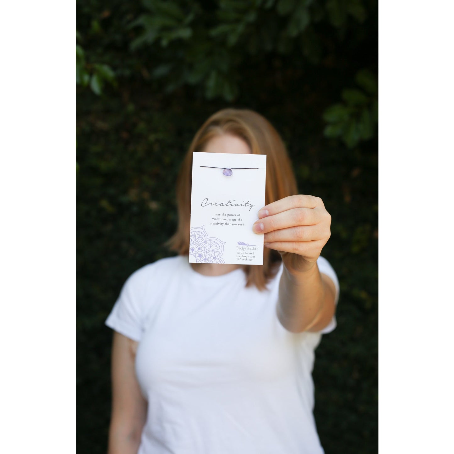A person holds up a "Creativity" necklace on a card with a faceted teardrop stone, obscuring their face.