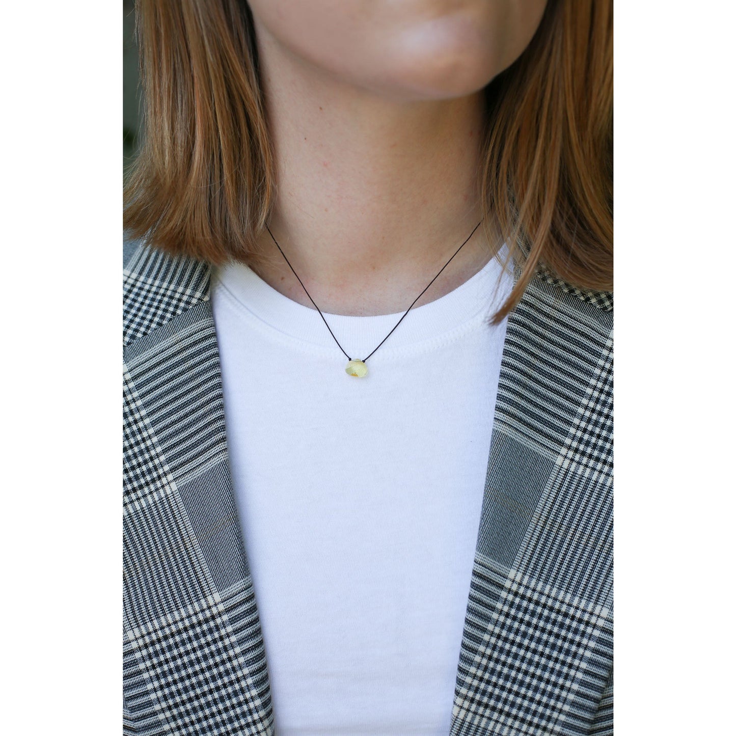 A person wears a black cord necklace with a yellow stone pendant, a white t-shirt, and a gray plaid blazer.
