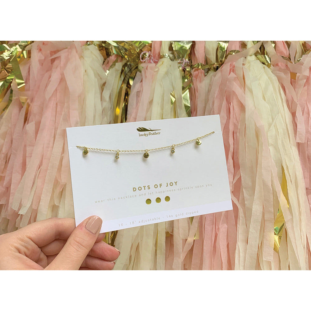 A hand holds a "Dots of Joy" necklace by Lucky Feather, displayed on a card, against a backdrop of pink, cream, and gold streamers.