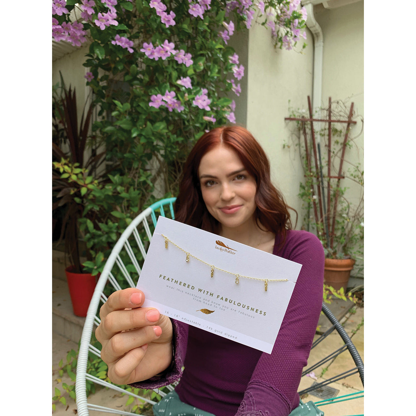 A woman holds up a card displaying a necklace, with purple flowers in the background.