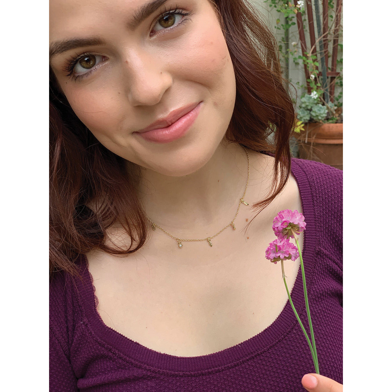 A woman smiles while holding two small pink flowers near her chest.