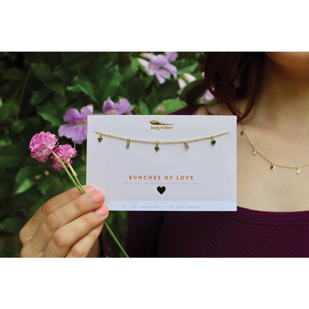 A woman displays a "Bunches of Love" necklace and packaging, holding pink flowers against a backdrop of foliage and purple flowers.