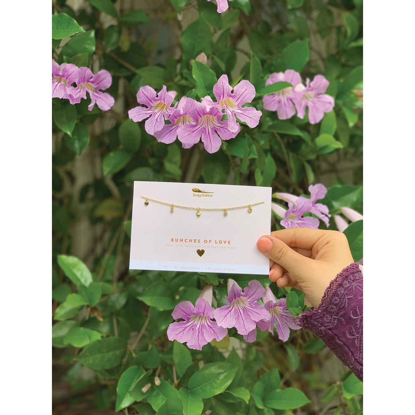 A hand holds a "Bunches of Love" necklace card against a backdrop of purple flowers and green leaves.