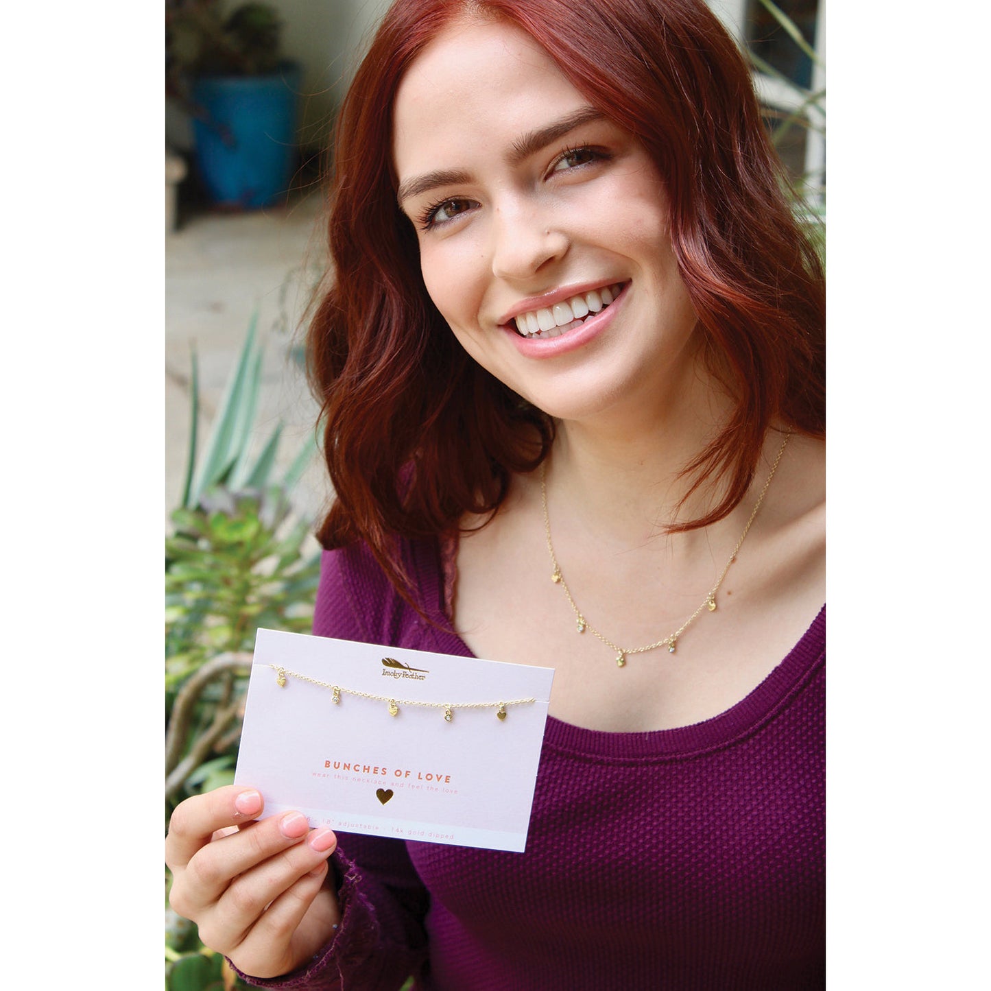 A smiling woman with red hair holds up a "Bunches of Love" necklace.