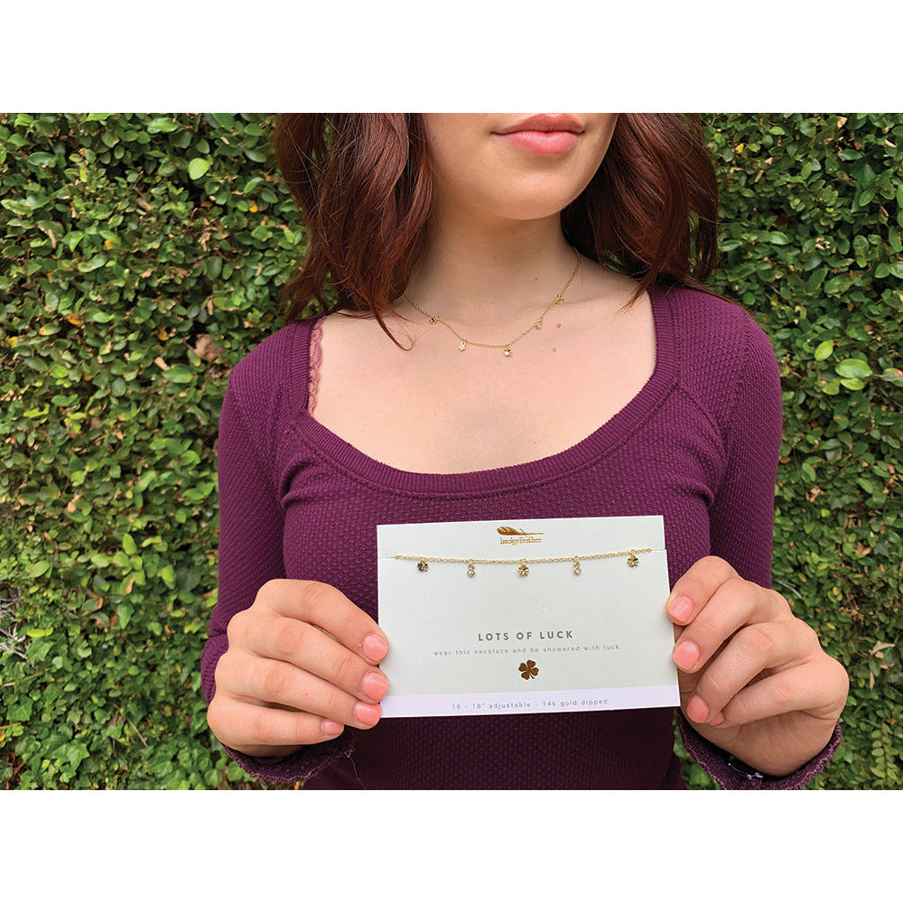 A woman wears a delicate gold necklace and holds a card displaying the same necklace labeled "Lots of Luck."