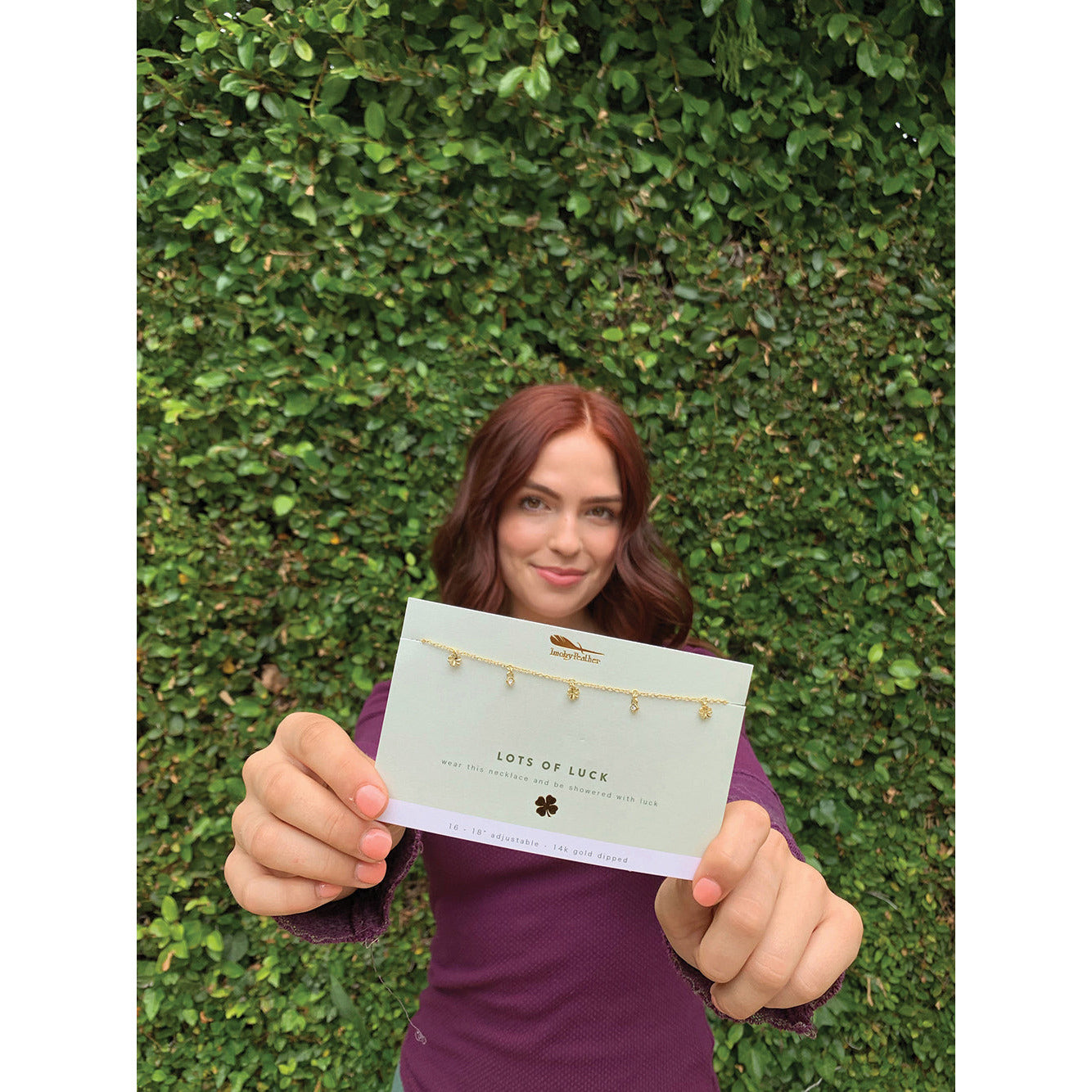 A woman holds up a "Lots of Luck" necklace package against a backdrop of greenery.
