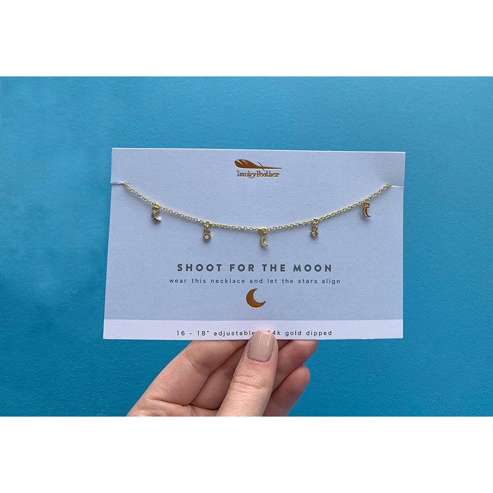 A hand holds a "Shoot for the Moon" necklace on its display card against a blue background.