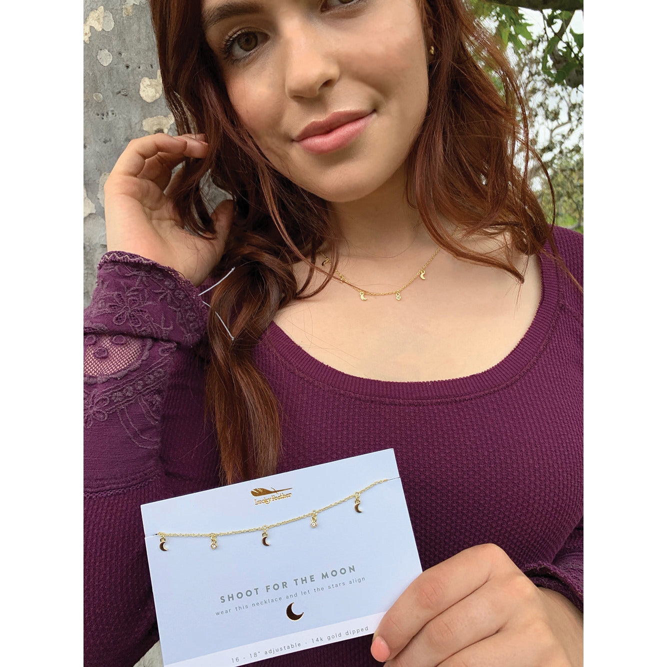A woman with auburn hair models a gold necklace with moon and star charms while holding the product packaging that reads "SHOOT FOR THE MOON".