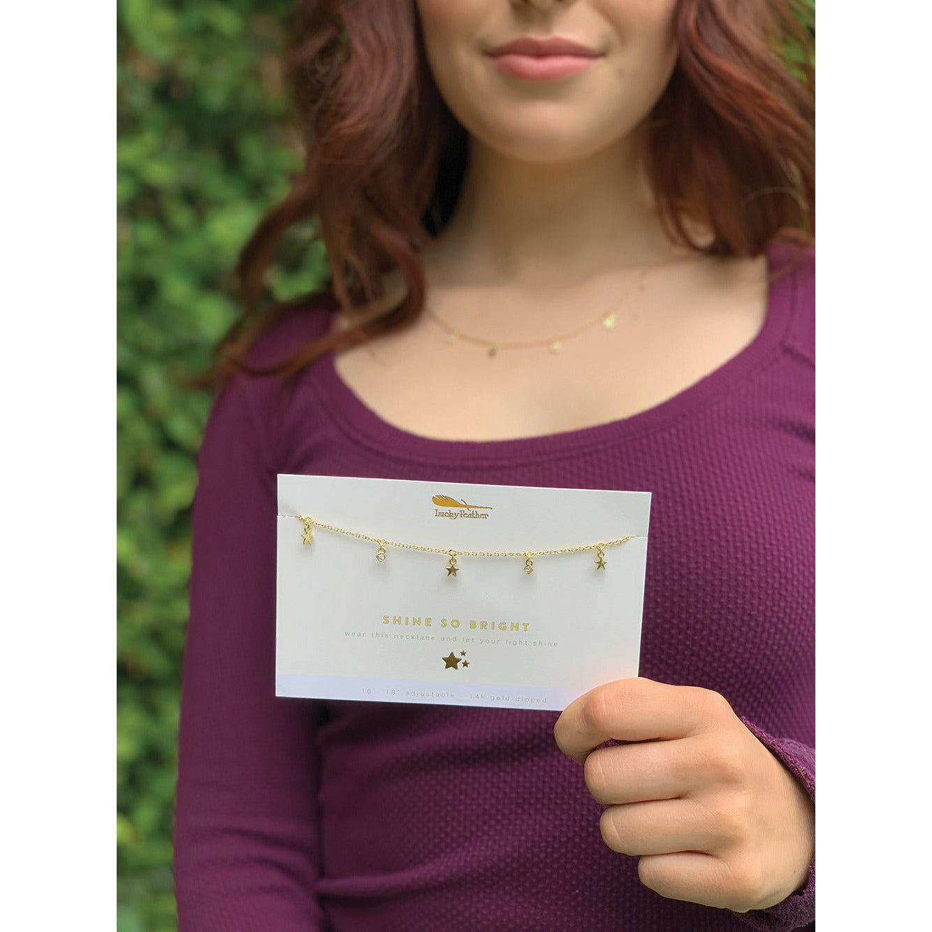 A woman holds a card displaying a "Shine So Bright" necklace by Irmaky Feather, featuring star and jewel pendants.