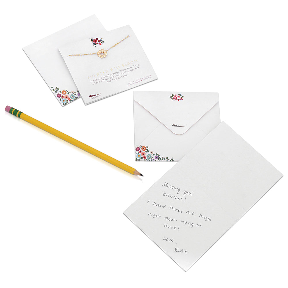A handwritten note and envelope with floral accents, alongside a yellow pencil and necklace on a card with a supportive message.