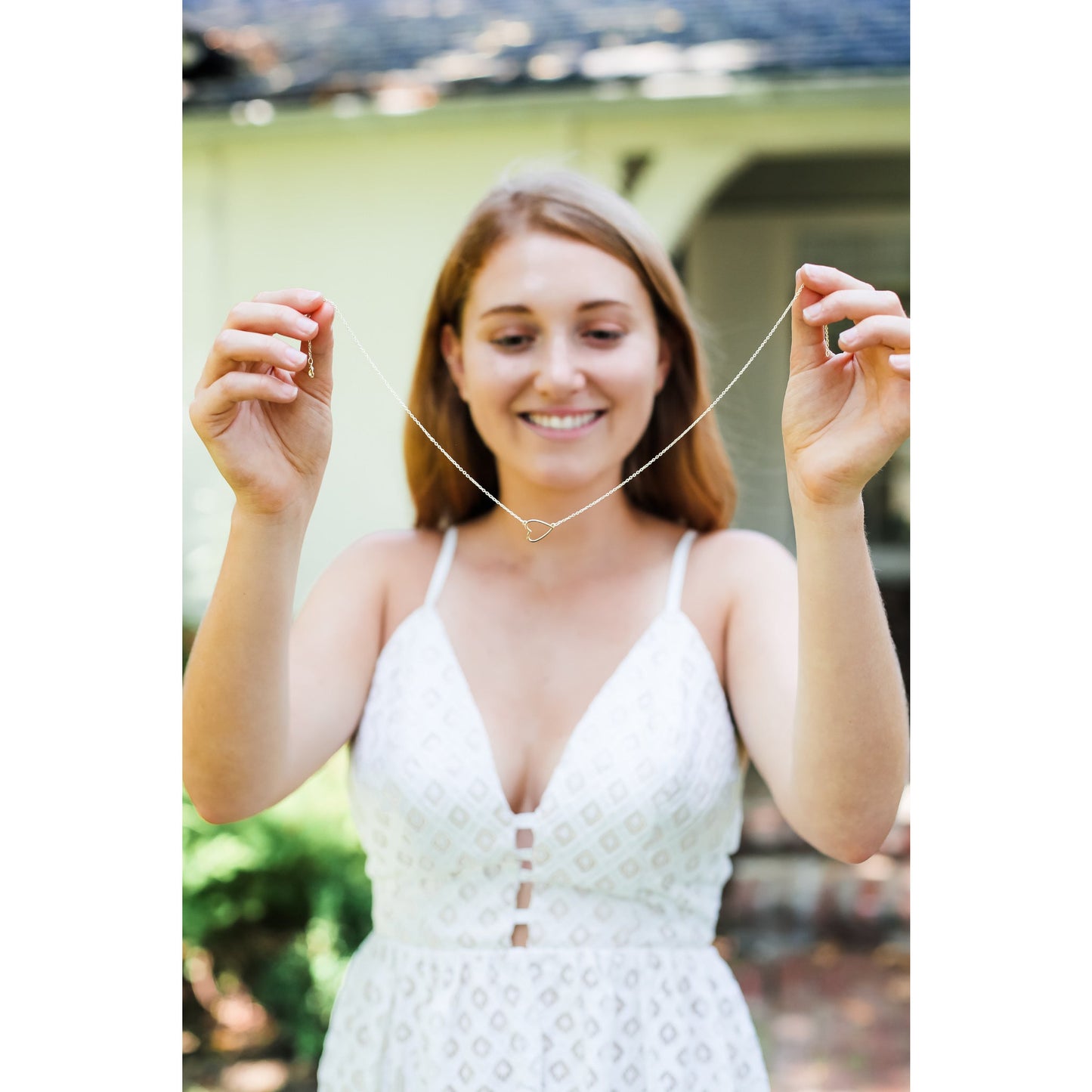 A woman in a white dress holds up a necklace with a heart pendant.