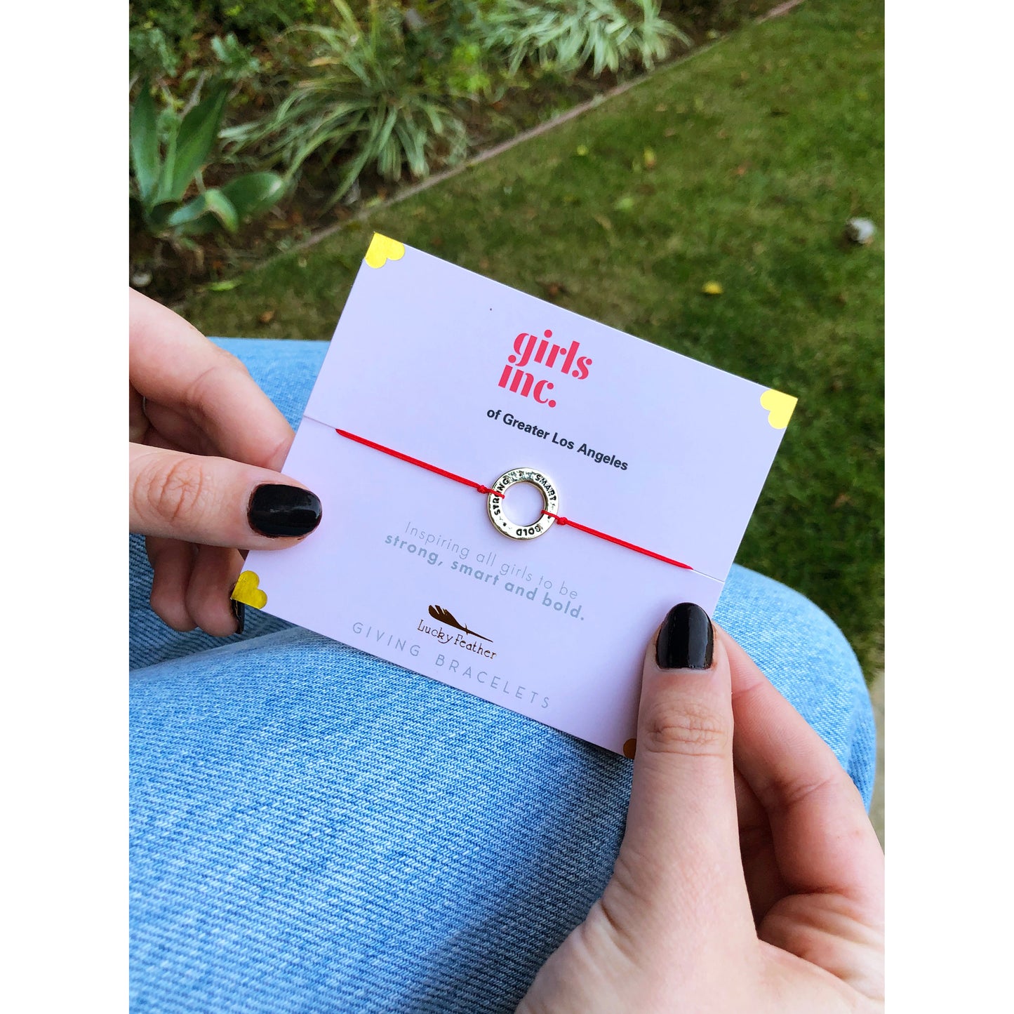 A person holding a card with a "girls inc." red string bracelet with a gold ring.
