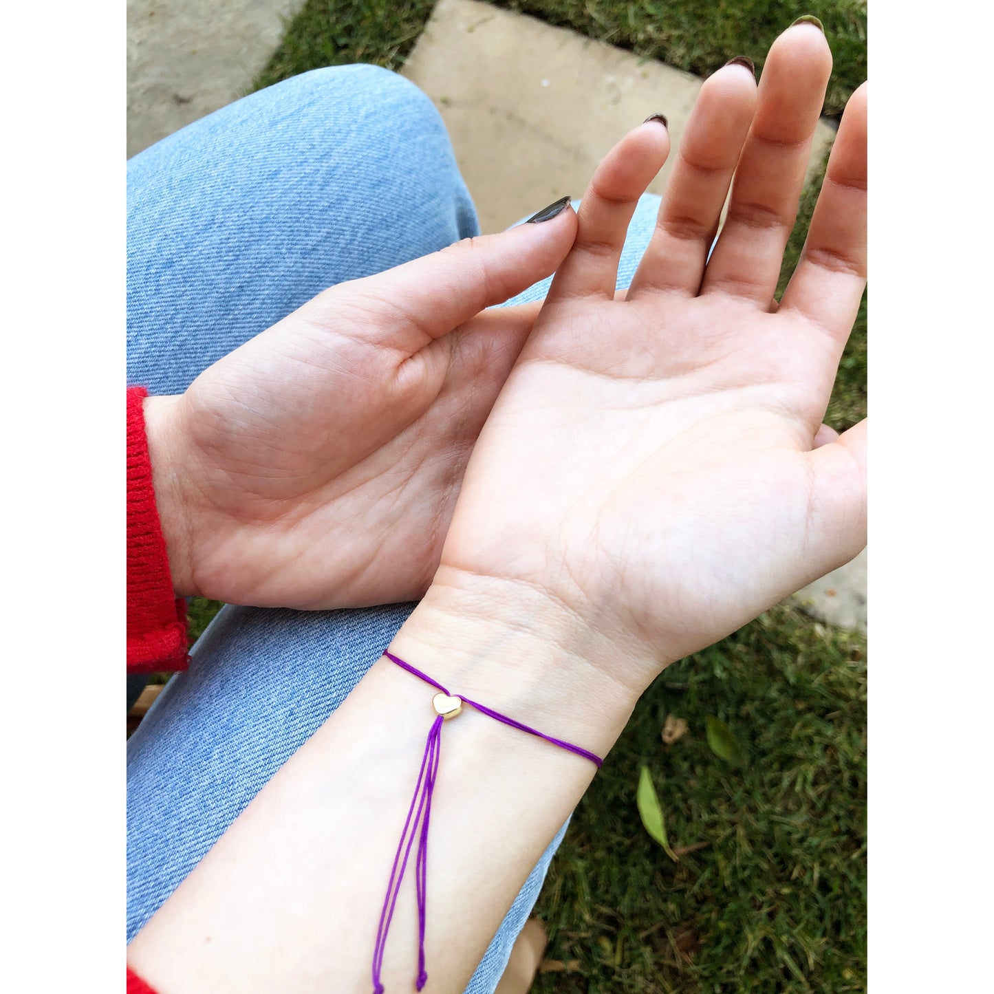 A person wearing a purple bracelet with a small gold heart charm is sitting in blue jeans.