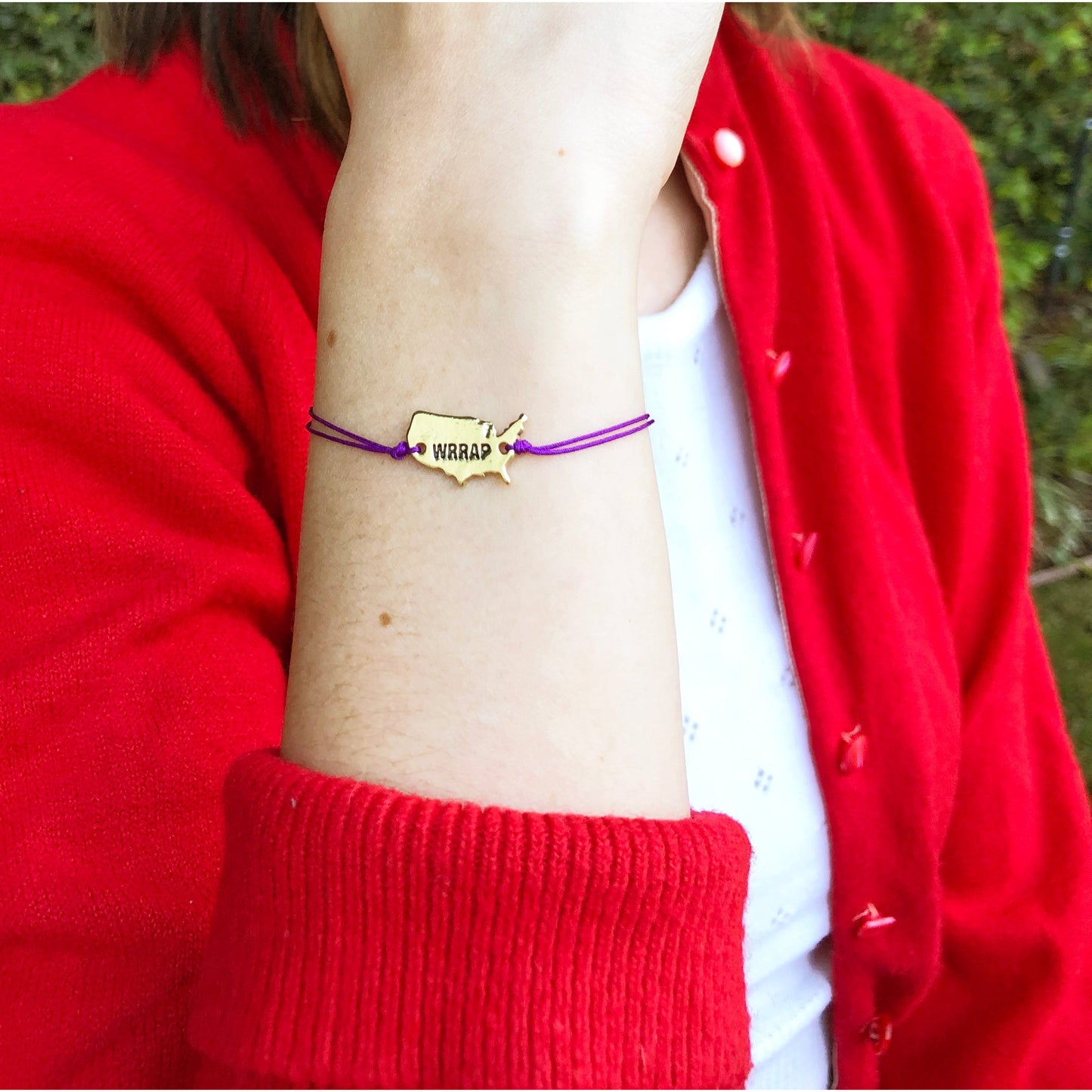 Person's wrist wearing a gold-colored bracelet with a United States shape and the word "WRRAP" on a purple string, with a red cardigan sleeve visible.