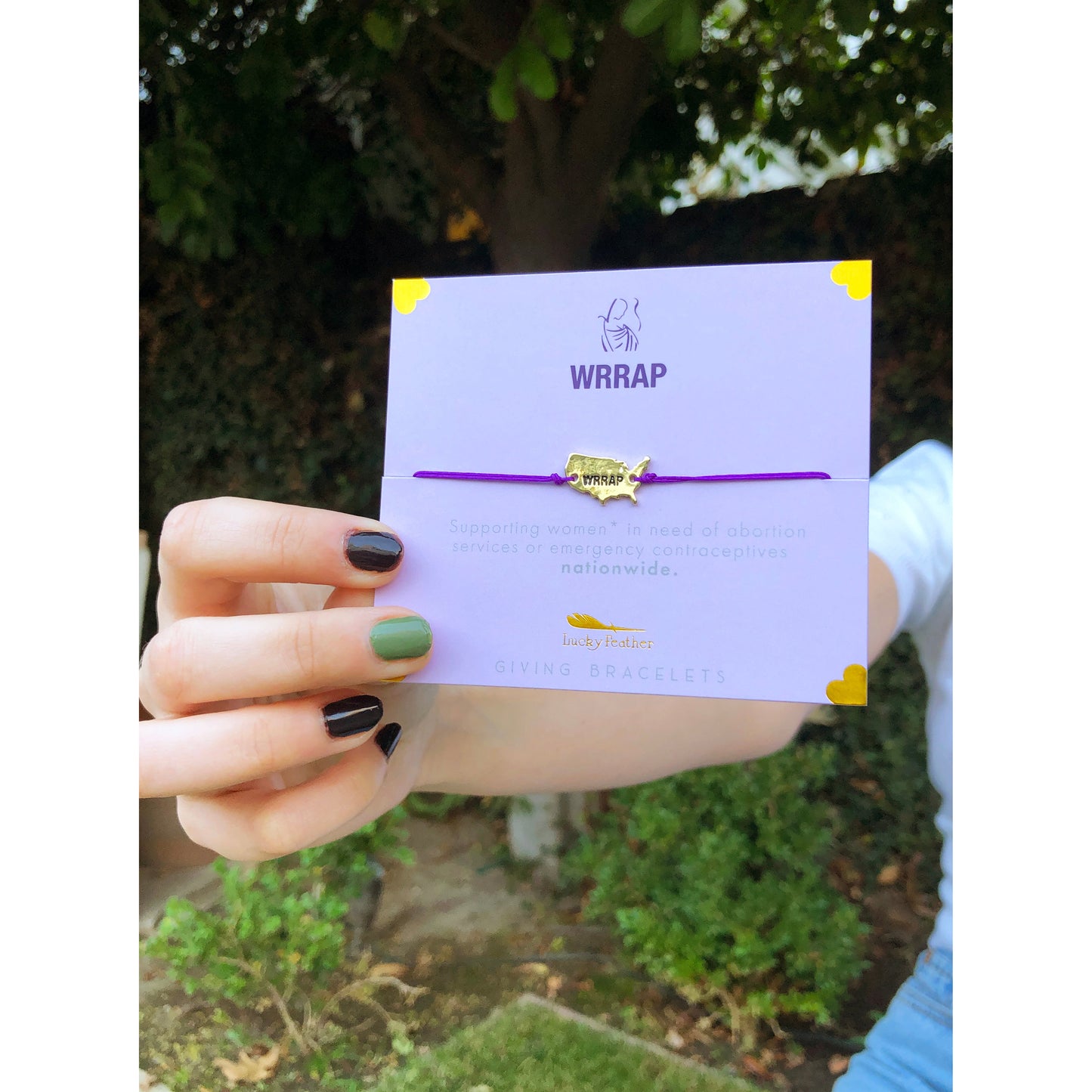 A hand holds a purple "Lucky Feather" giving bracelet on a lavender card with the WRRAP logo.