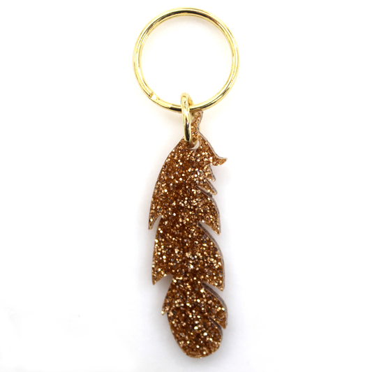 A gold-colored glittery feather charm hangs from a gold key ring.