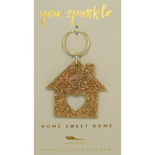 A golden glittery house-shaped keychain with a heart-shaped cutout hangs from a tag that reads 'you sparkle' and 'home sweet home.'