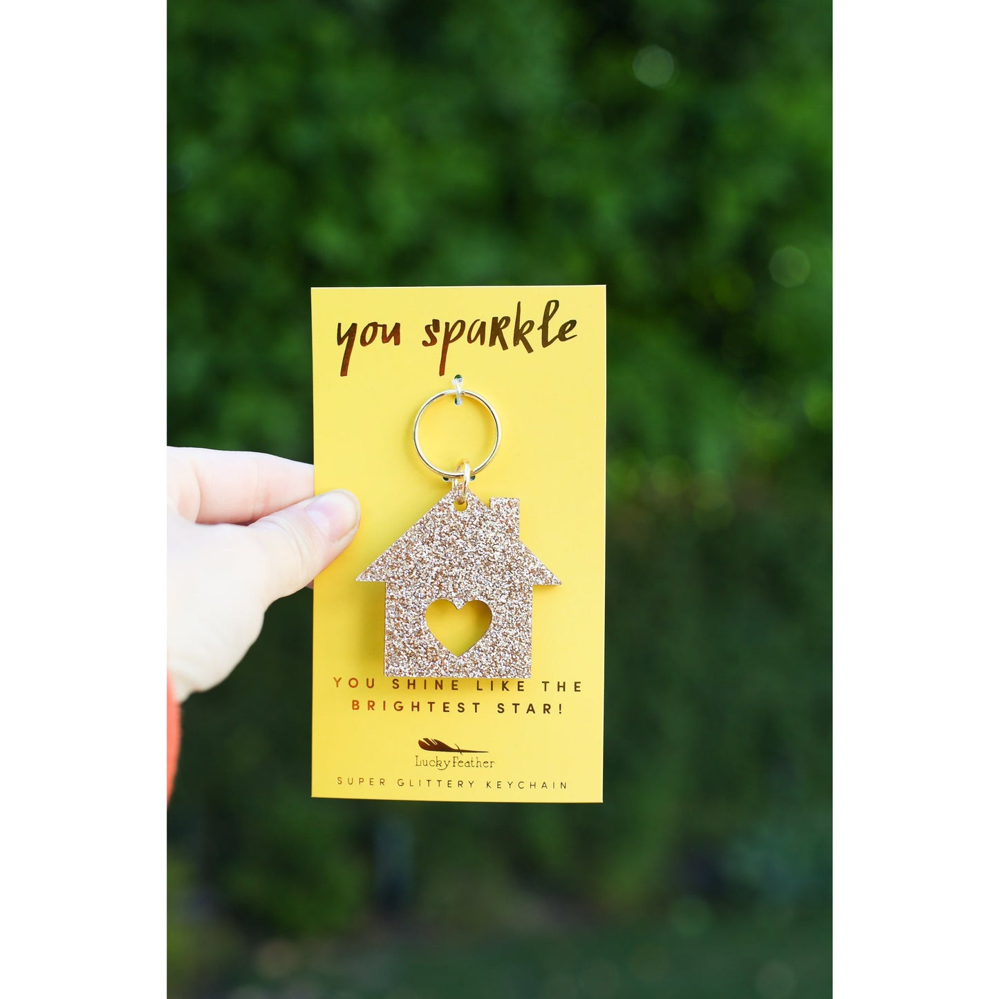 A hand holds up a 'Lucky Feather' brand super glittery house-shaped keychain with a heart cutout on a yellow card that reads, 'you sparkle' and 'YOU SHINE LIKE THE BRIGHTEST STAR!'.
