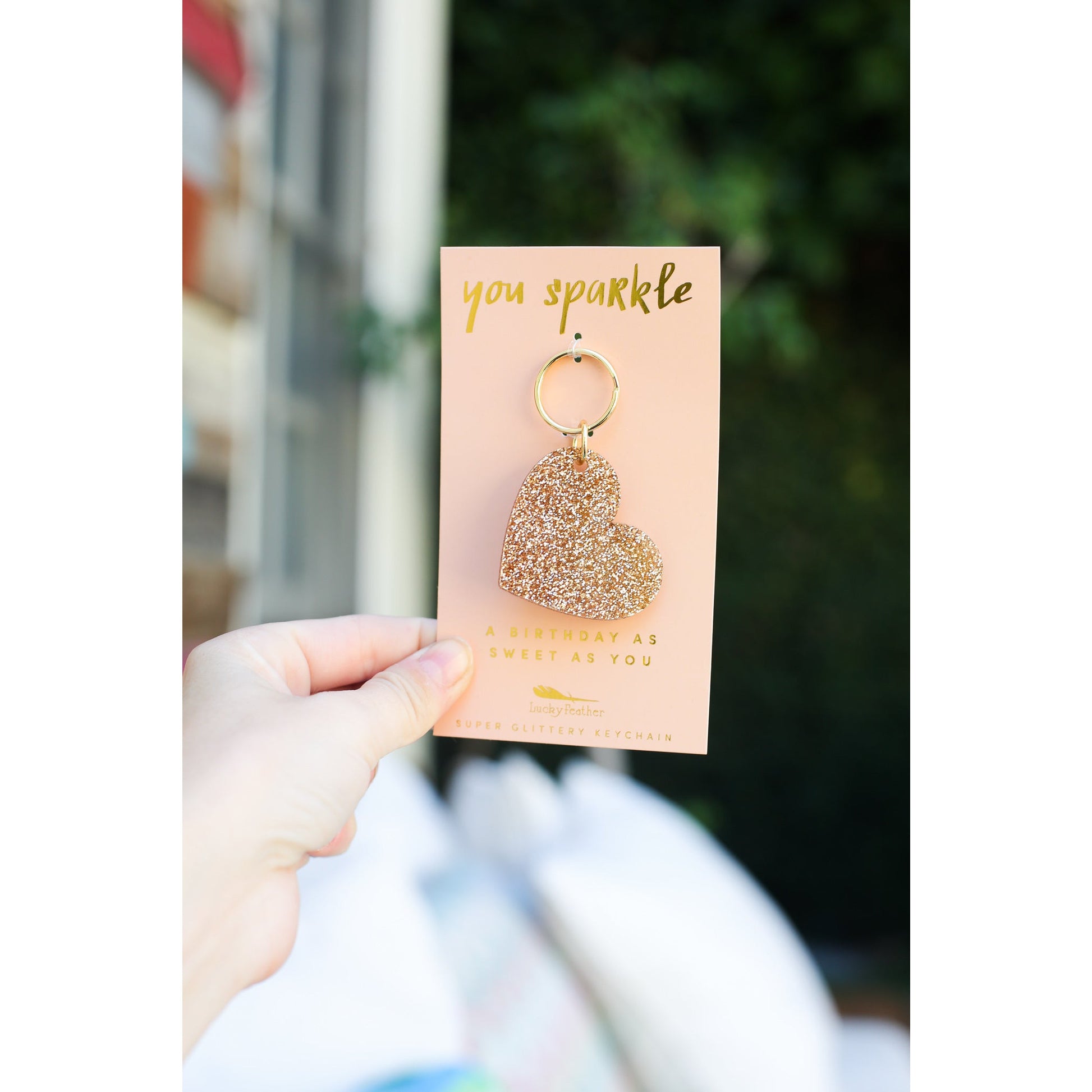A hand holds a peach-colored card with a gold glitter heart-shaped keychain attached.