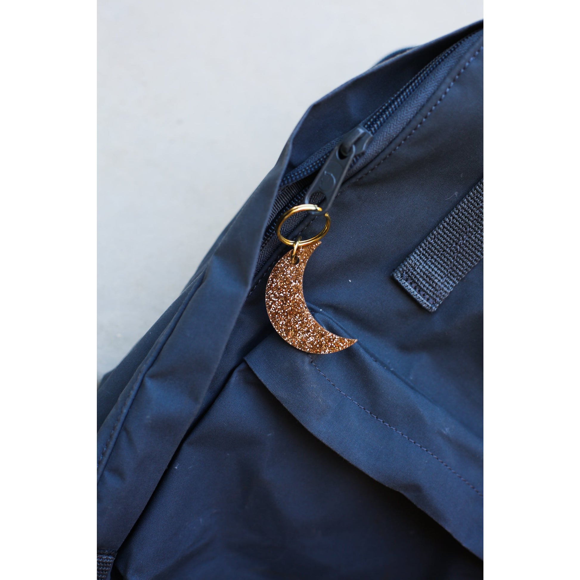 A glittery crescent moon keychain hangs from the zipper of a navy blue backpack.