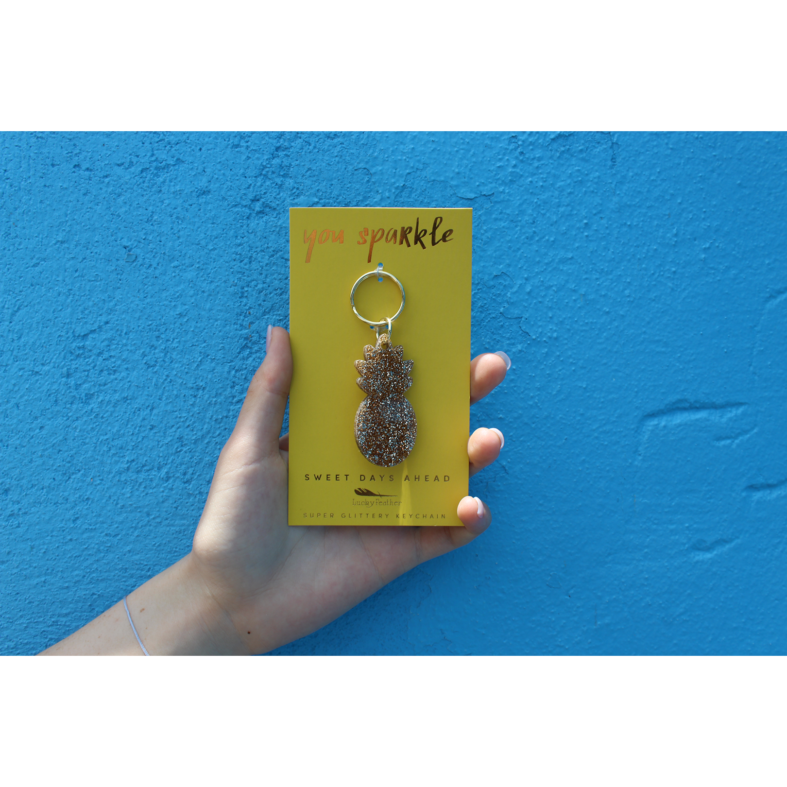 A hand holds a yellow card with a glittery pineapple keychain against a textured blue background.