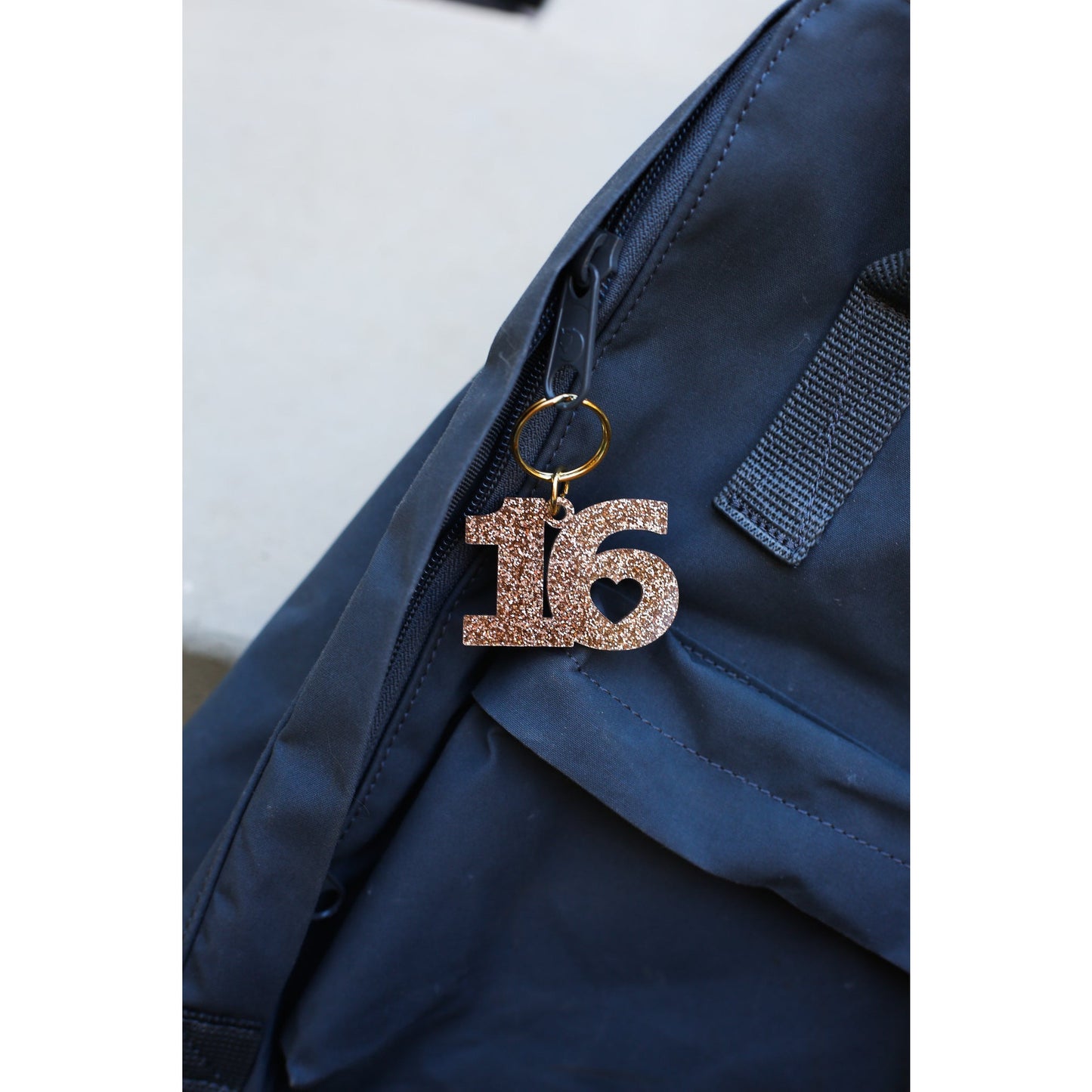 A sparkly, rose gold number 16 keychain with a heart detail hangs from a blue backpack.