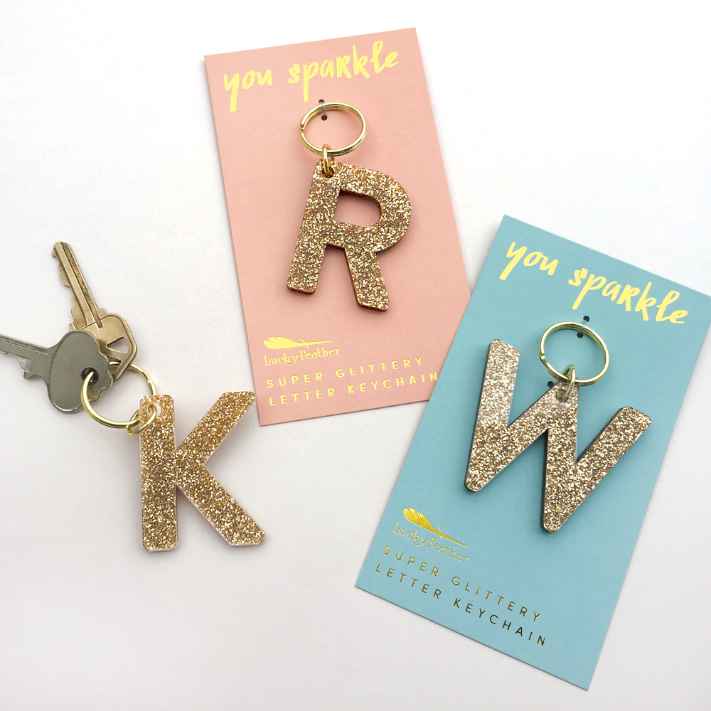 Three gold glitter letter keychains are displayed on their retail cards.
